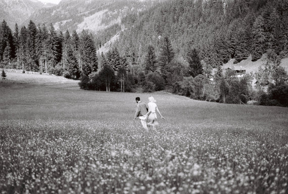 A figure walking through a wide meadow below a dark treeline